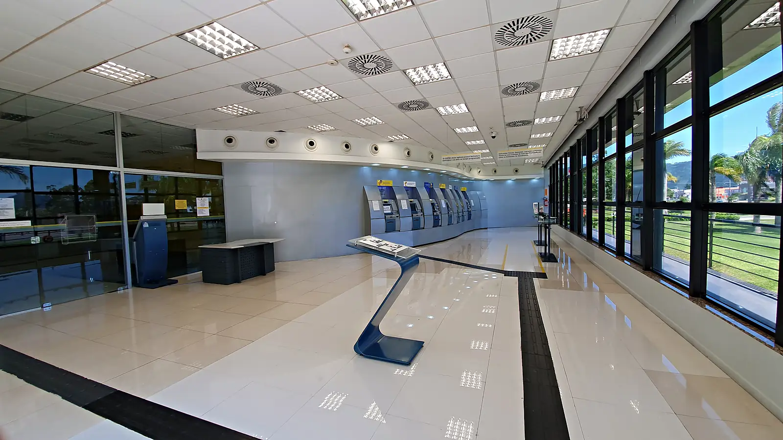 Sala de autoatendimento de uma agência do Banco do Brasil, com a fila de caixas eletrônicos ao fundo, piso claro e janelas de vidro à direita.