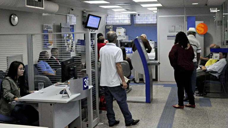 Salão de atendimento de uma agência da Caixa Econômica Federal, com uma funcionária na mesa de atendimento 36, clientes em pé e sentados na espera e o painel de senhas no alto.