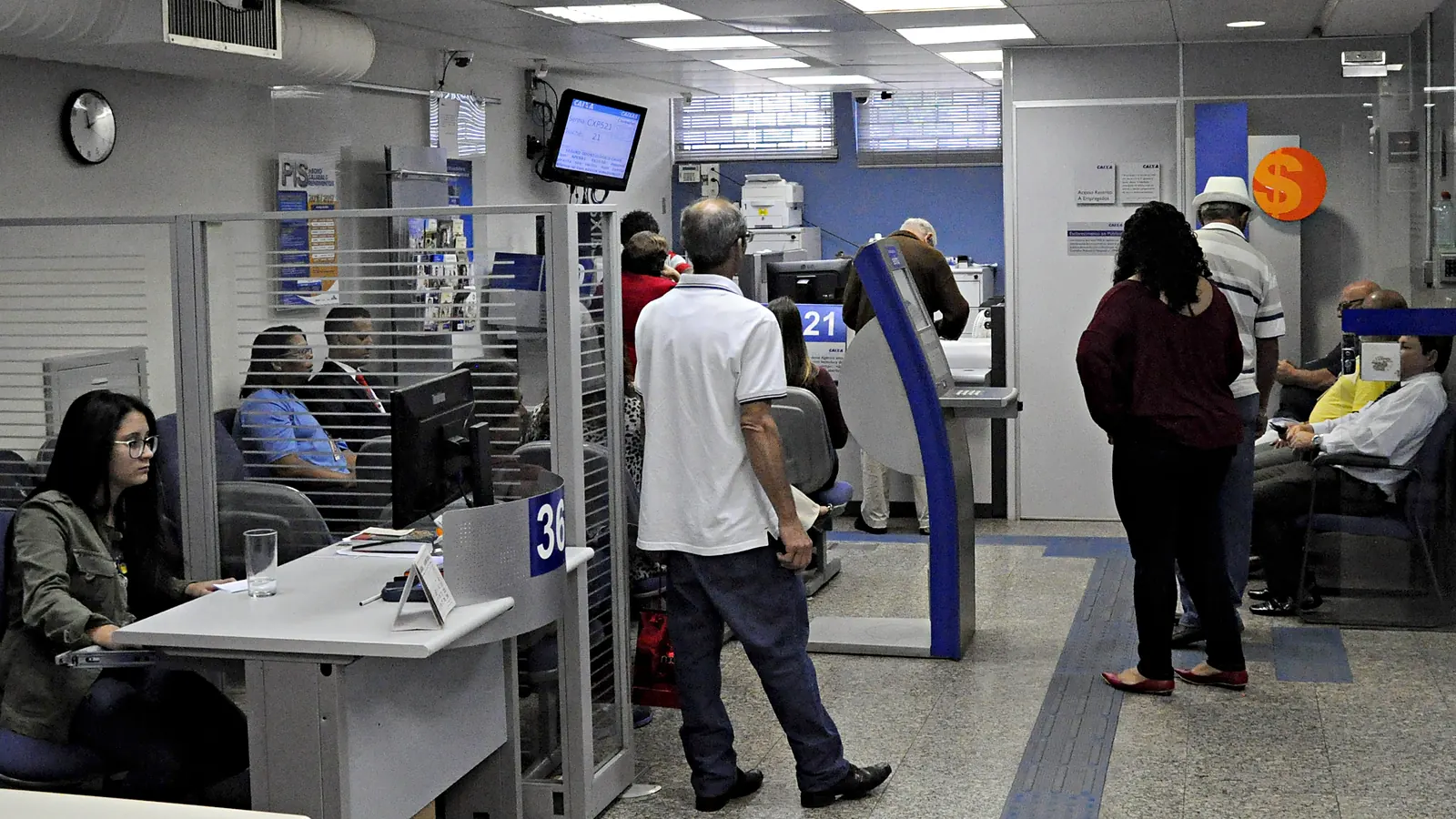 Salão de atendimento de uma agência da Caixa Econômica Federal, com uma funcionária na mesa de atendimento 36, clientes em pé e sentados na espera e o painel de senhas no alto.