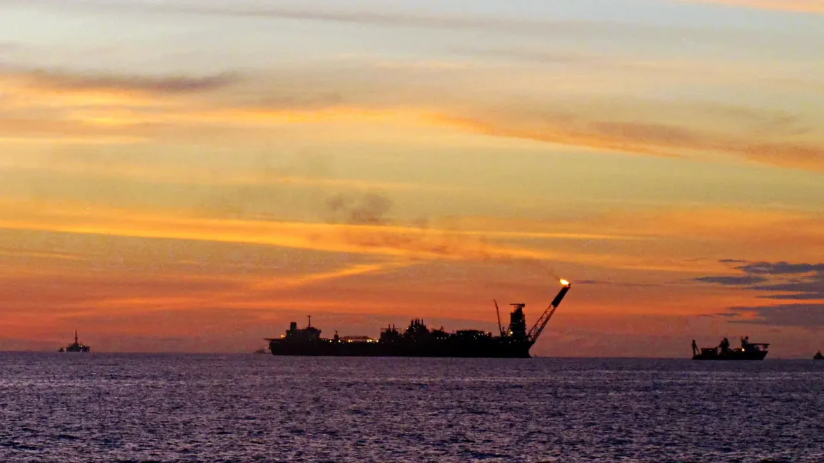 Navio-plataforma de produção de petróleo na Bacia de Campos ao entardecer, em silhueta contra o céu laranja, com a chama do queimador acesa e barcos de apoio no horizonte.