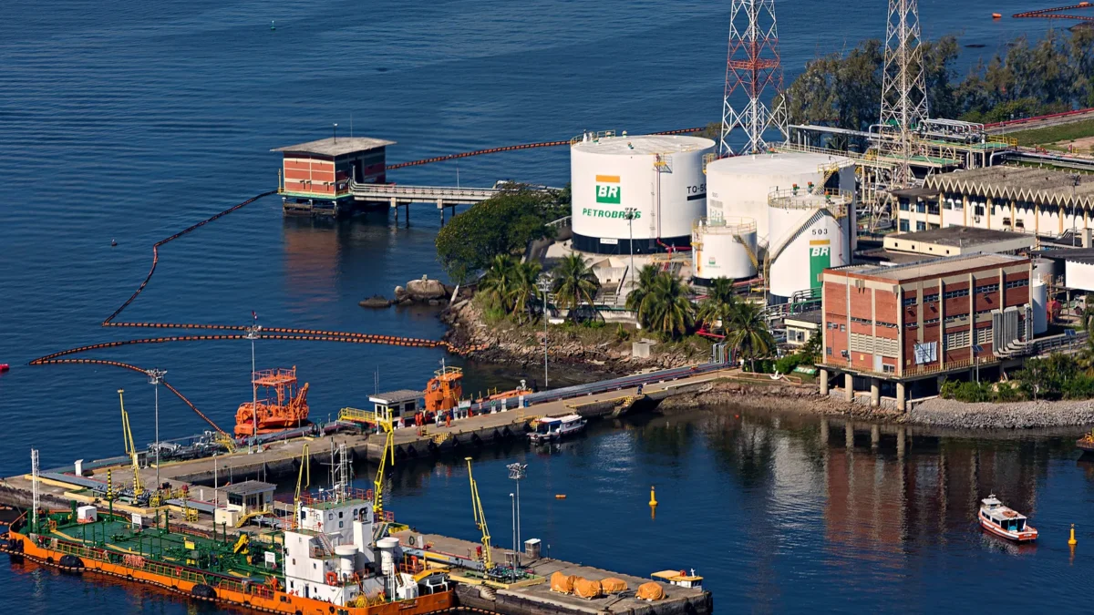 Vista aérea do terminal da Transpetro na Ilha d'Água, no Rio de Janeiro, com tanques brancos de armazenamento, píer com uma embarcação atracada e boias de contenção na baía.