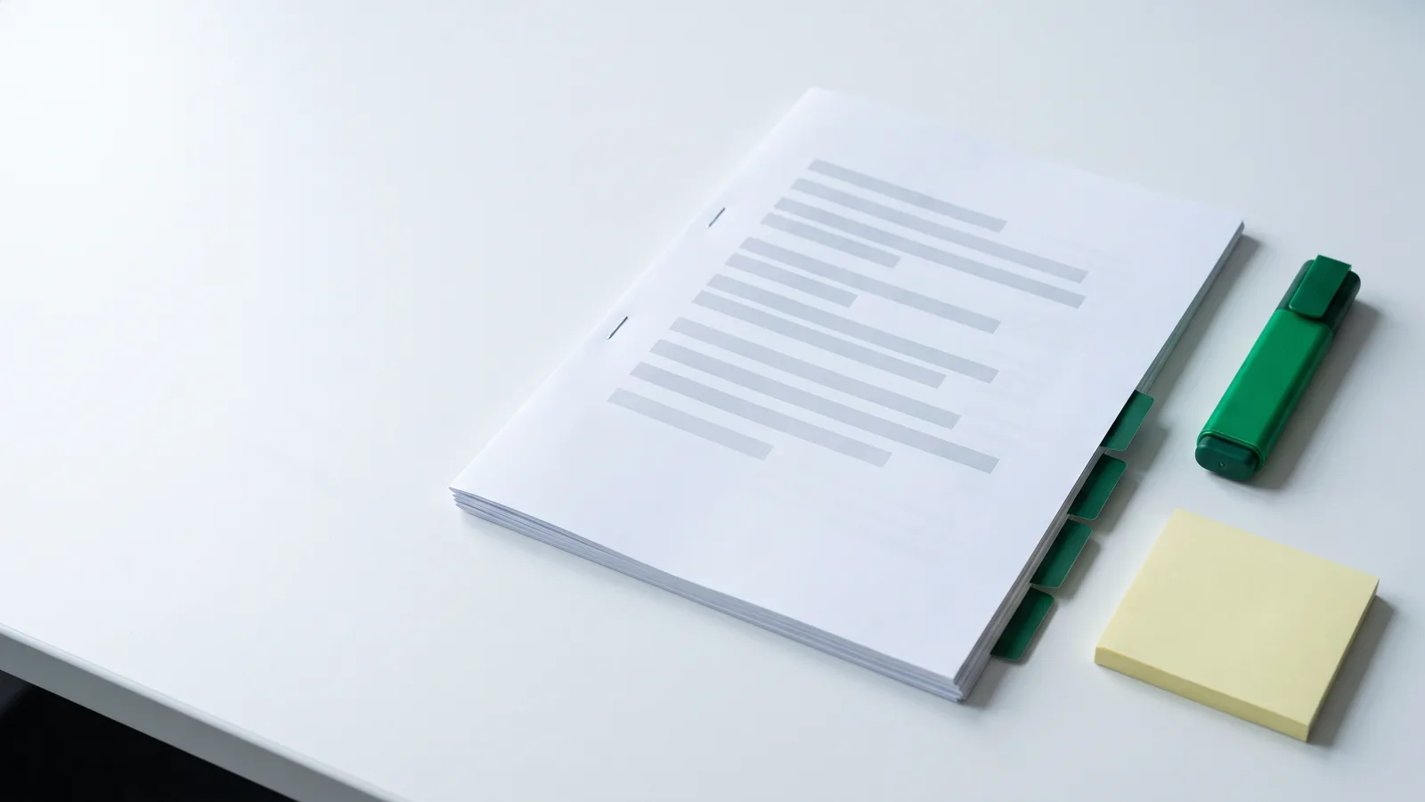 Edital de concurso impresso e grampeado sobre uma mesa branca, com quatro divisórias verdes marcando os anexos, um marca-texto verde e um bloco de notas amarelo ao lado.