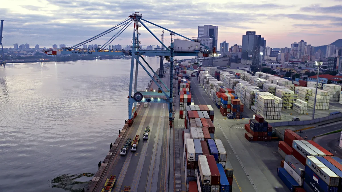 Vista do alto do terminal de contêineres do Porto de Itajaí, com um portêiner azul no cais, pilhas de contêineres no pátio, caminhões de terminal e o rio Itajaí-Açu à esquerda, com prédios da cidade ao fundo.