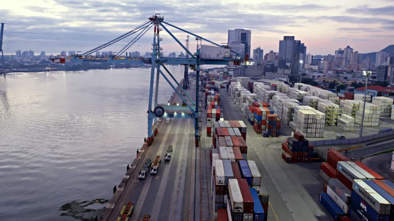 Vista do alto do terminal de contêineres do Porto de Itajaí, com um portêiner azul no cais, pilhas de contêineres no pátio, caminhões de terminal e o rio Itajaí-Açu à esquerda, com prédios da cidade ao fundo.