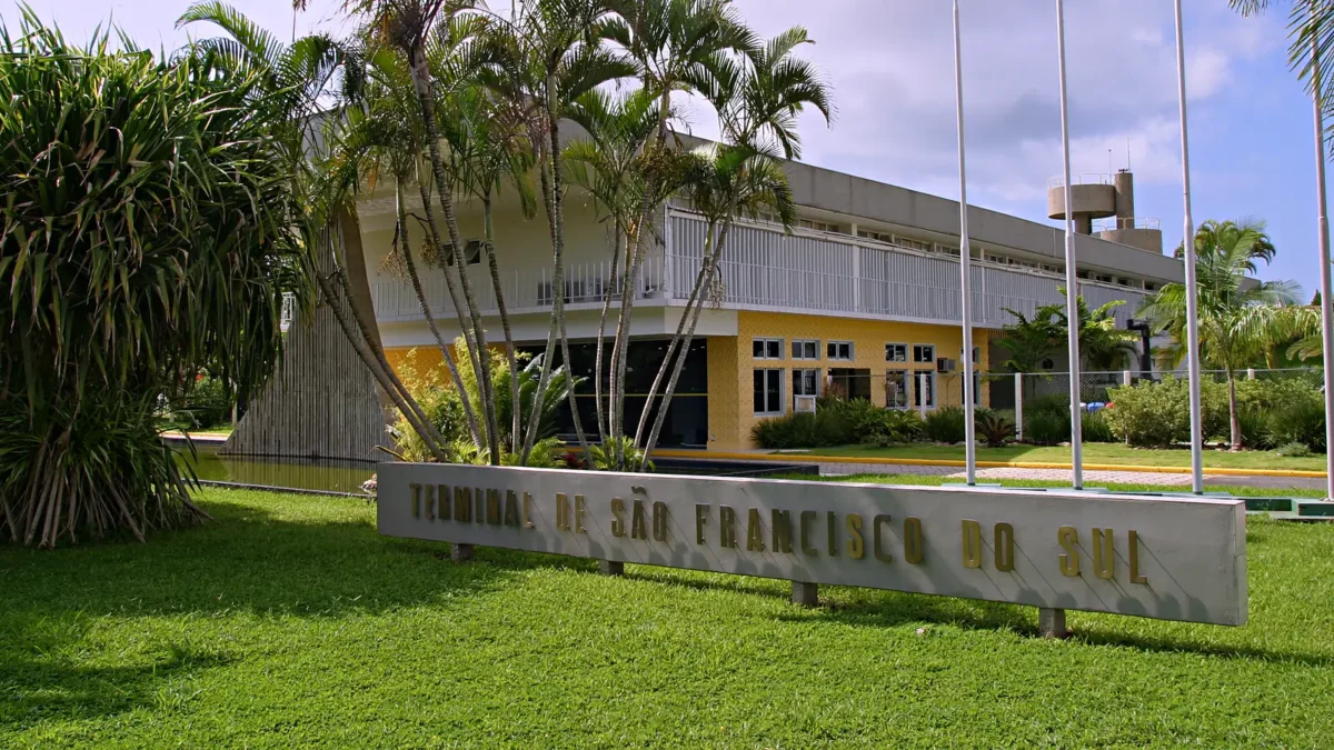 Entrada do Terminal de São Francisco do Sul, da Transpetro, com a placa do terminal sobre o gramado, palmeiras e o prédio administrativo de fachada amarela ao fundo.