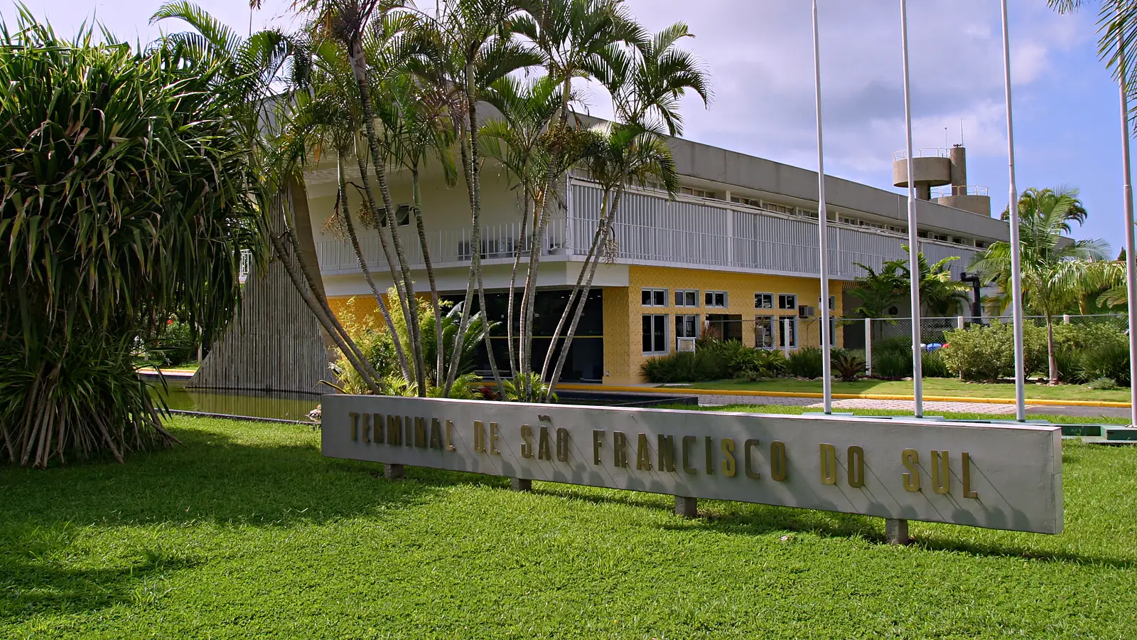 Entrada do Terminal de São Francisco do Sul, da Transpetro, com a placa do terminal sobre o gramado, palmeiras e o prédio administrativo de fachada amarela ao fundo.