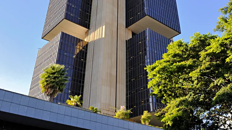 Edifício-sede do Banco Central do Brasil, em Brasília, visto de baixo, com a torre de vidro escuro e colunas bege sob céu azul e árvores no primeiro plano.