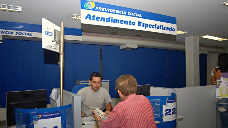 Servidor do INSS atende um segurado no guichê de uma agência da Previdência Social, sob a placa de Atendimento Especializado, enquanto o segurado mostra um documento.